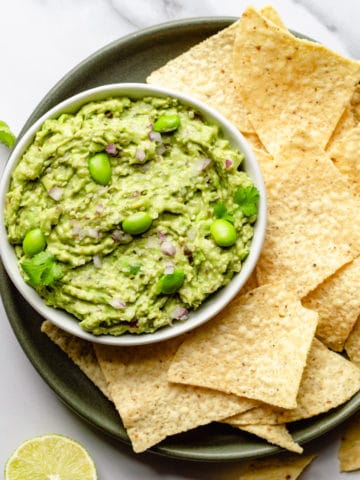 edamame guacamole in a white bowl on a green plate filled with tortilla chips