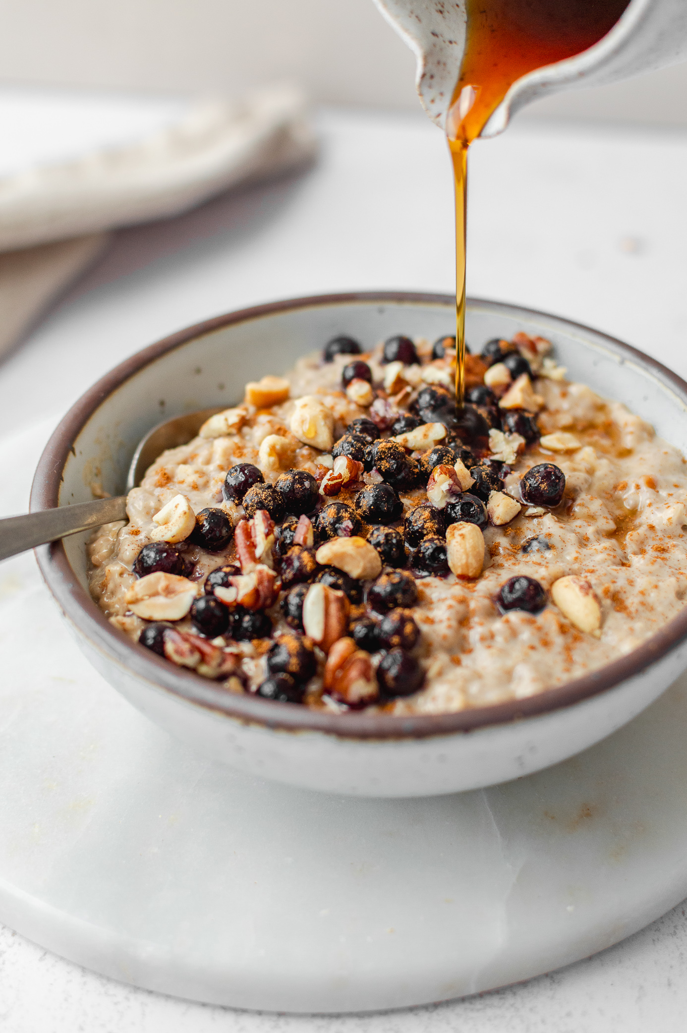 Maple syrup being poured over creamy peanut butter oatmeal with blueberries and chopped peanuts