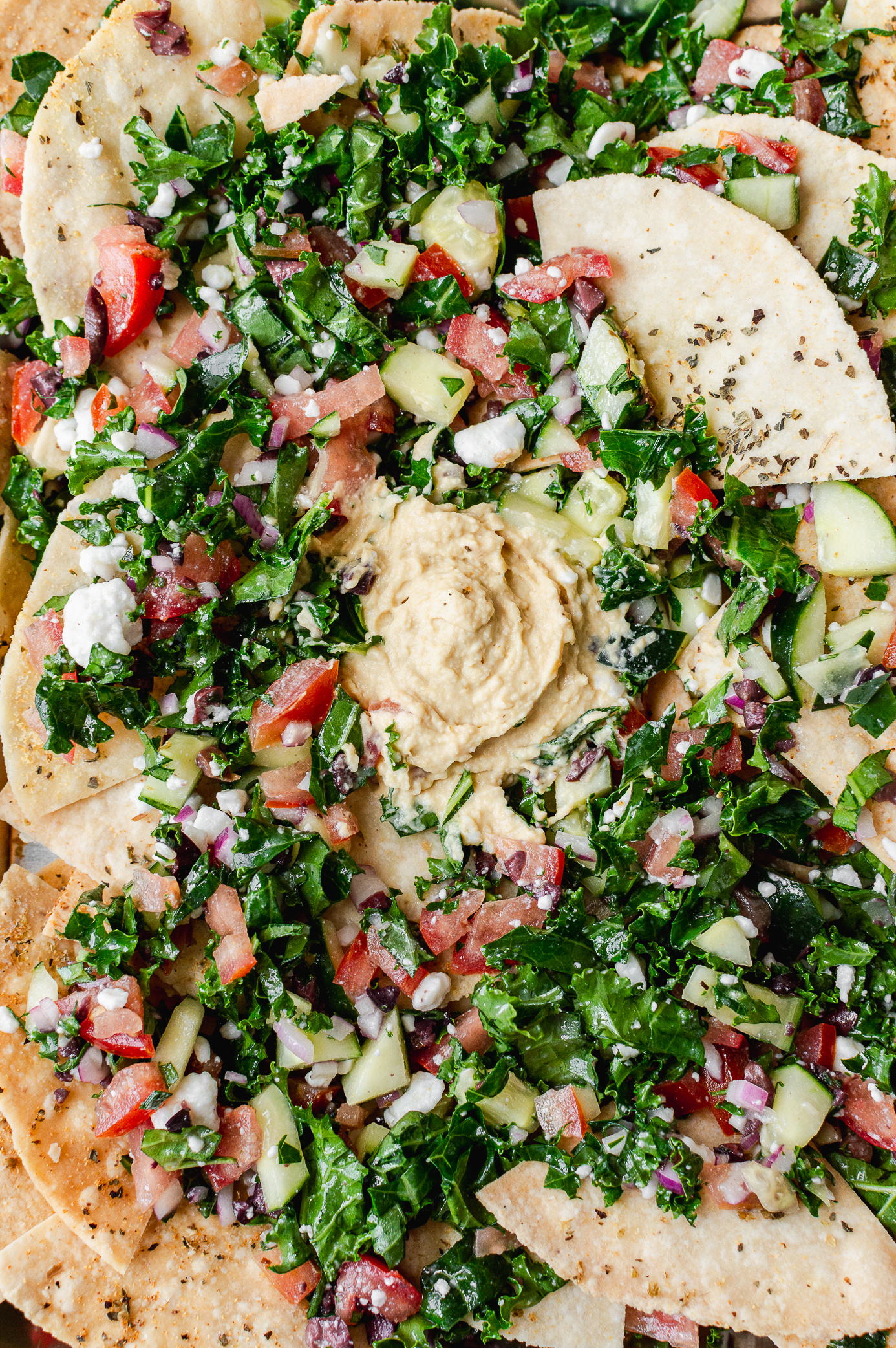 Platter of vegan Mediterranean nachos with hummus spread in the center and loaded pita chips around it.