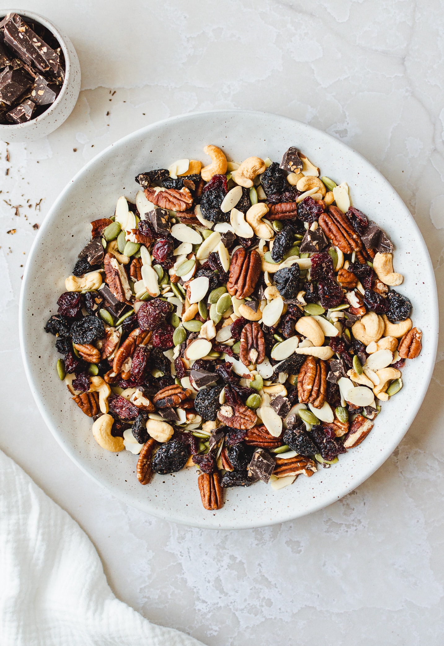 Styled homemade vegan trail mix scene with a ceramic bowl, soft linen, and scattered chocolate chunks.