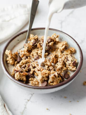 Chocolate chip cookie dough granola clusters in a ceramic bowl with milk being poured over the top.